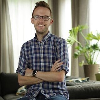 A man in a plaid shirt and glasses, smiling at the camera with arms crossed, stands in the living room of a short-term rental property.