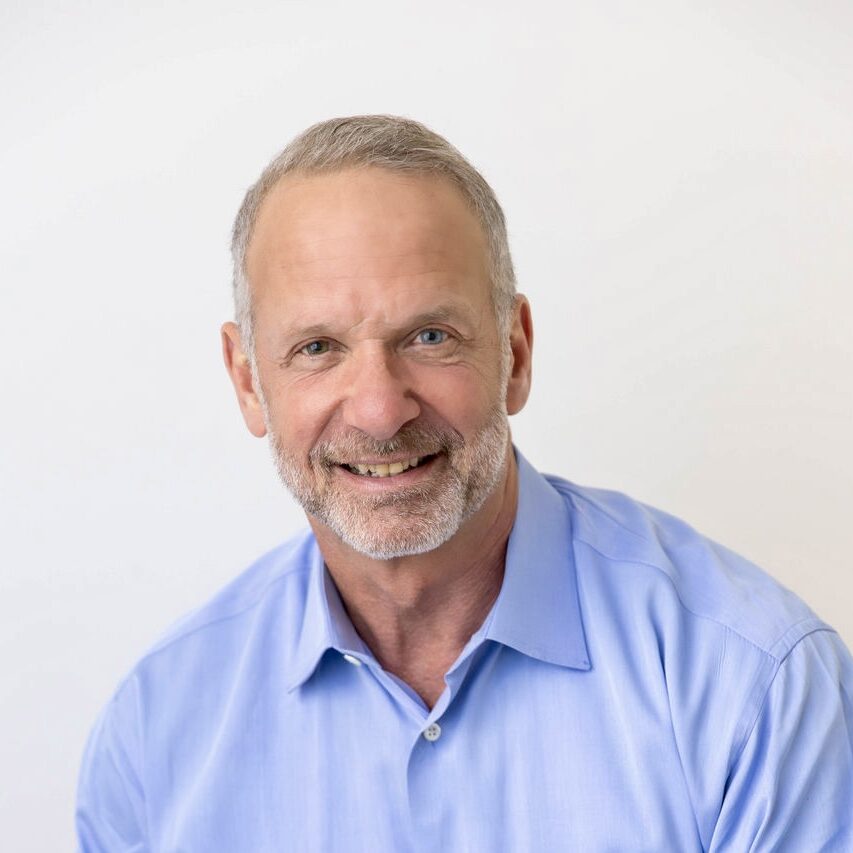 A man with short gray hair and a beard is smiling while wearing a light blue collared shirt against a plain white background.