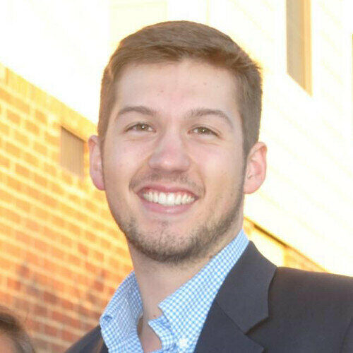 A person smiling outdoors in front of a brick building.