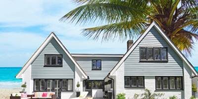 A modern two-story house with light grey and white exterior is situated by the beach, with a large palm tree and blue ocean in the background.