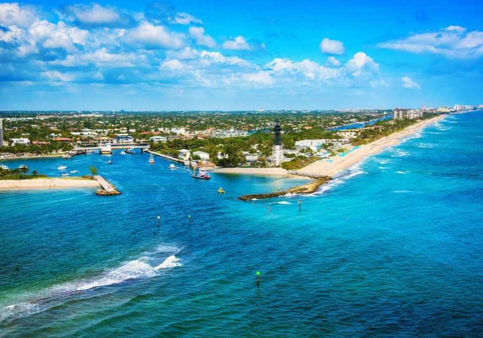 Aerial view of a lush coastline with a sandy beach, boats sailing on vivid blue waters, a bridge crossing a narrow water channel, and a distant cityscape under a clear sky, ideal for vacation