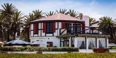 Two-story house with red shutters, white exterior, and a partially covered porch. The house is surrounded by palm trees and a clear sky.