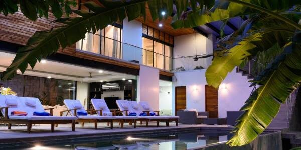 Outdoor view of a modern house with a swimming pool, surrounded by lounge chairs with white cushions and a banana tree in the foreground. The house is well-lit and has large windows and wooden accents.