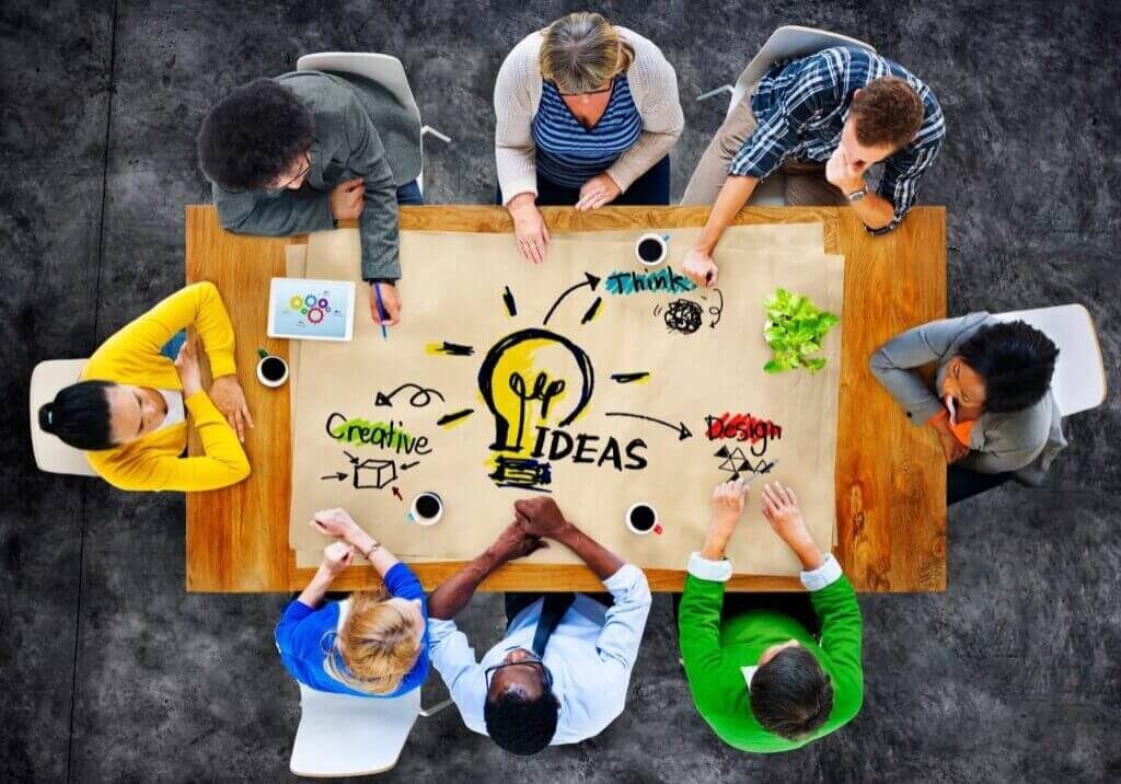 A diverse group of people seated around a table brainstorming, with the word "IDEAS" and drawings related to creativity, design, and strategy sketched on a large paper in the center.