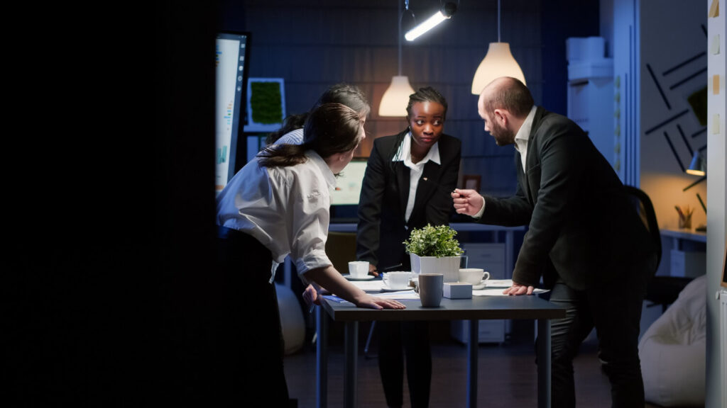 Three professionals, one man and two women, stand around a meeting table discussing papers in a dimly lit office of a property management business.