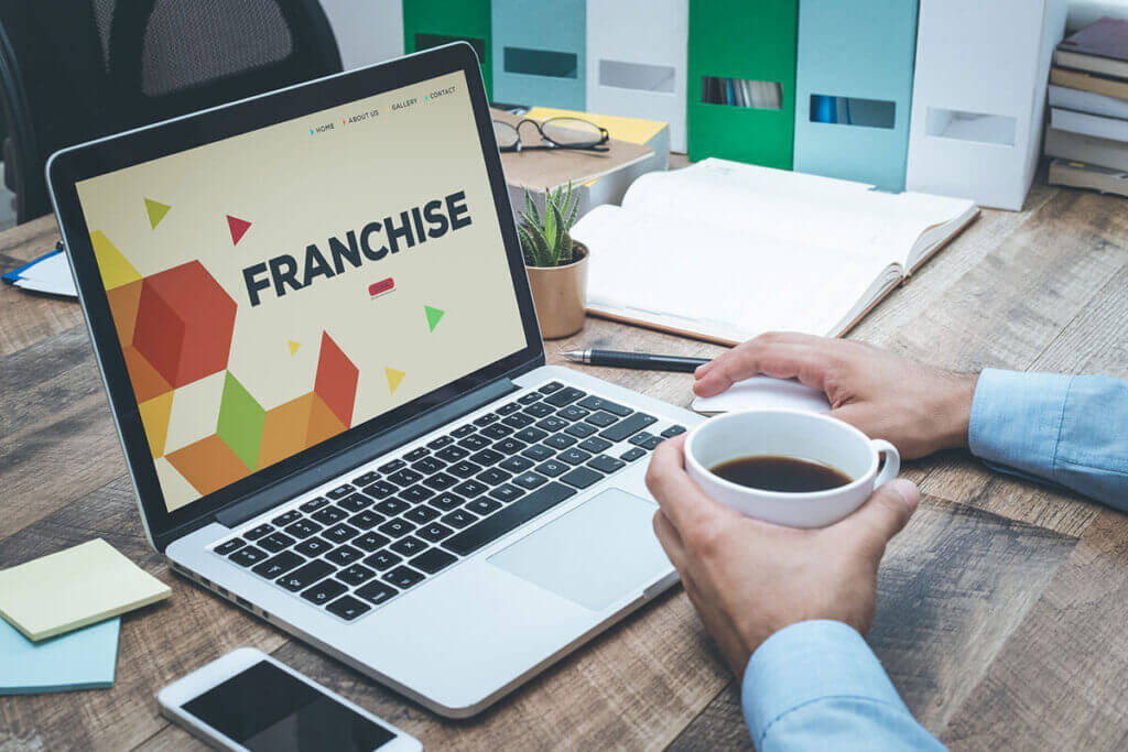 A person holds a cup of coffee while viewing a laptop screen displaying the word "FRANCHISE" in an office setting.